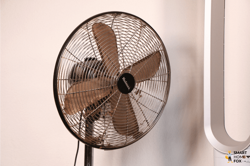 За защитной решеткой старого вентилятора с лопастями находятся комки пыли